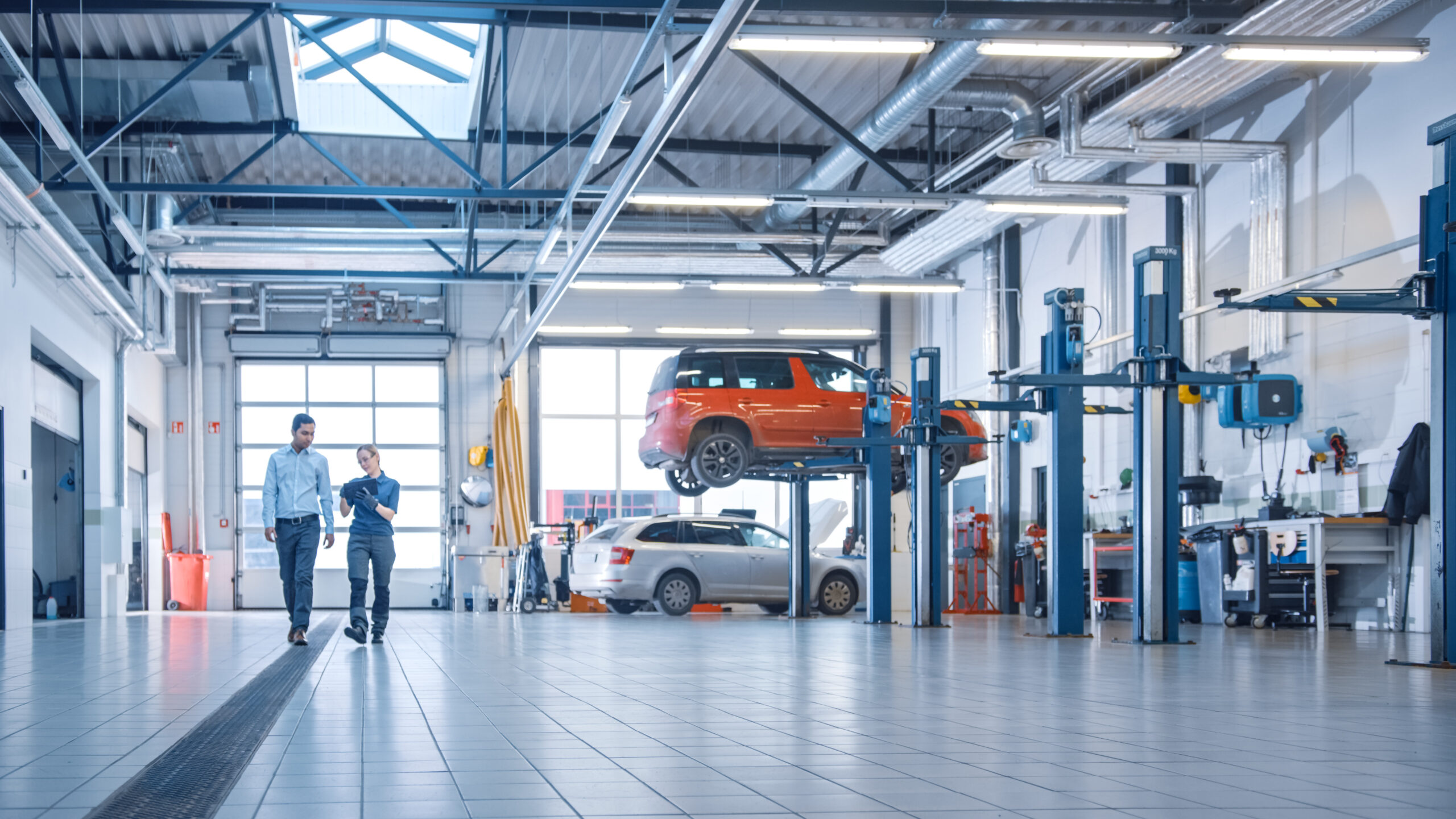 Female Mechanic Checks Diagnostics Results on a Tablet Computer and Explains a Vehicle Breakdown to a Manager. Car Service Employees Talk while Walking in a Garage. Modern Clean Workshop.